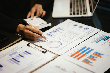 Young Asian businesswoman pointing pen with graph and chart report and using laptop computer to analyze data.