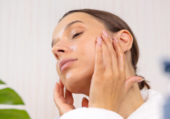 woman applying face sheet mask at home, cream or serum and massaging skin with roller.girl doing beauty routine at home. double side mirror and cream jar on table,monstera plant leaf in background.