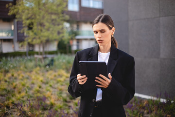 Fototapeta premium Serious, elegant businesswoman standing on the street hold tablet. Success coming with hard work. Young proud woman with low ponytail wear suit on a street watch at tablet.