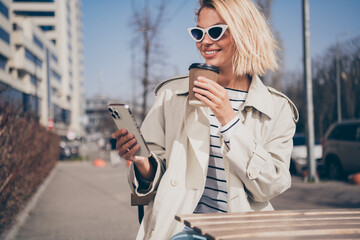 Photo of pretty young blonde woman sit table hold device drink coffee wear stylish trench coat walk outside urban city streets