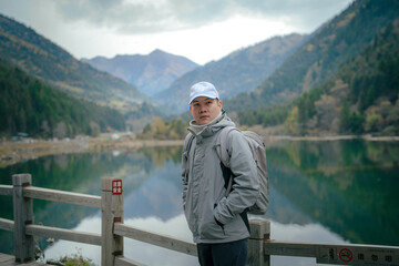 Fototapeta premium Hiker man wearing jacket and backpack standing by the mountain lake and looking at camera, Dagu Glacier, Chengdu, China. 
