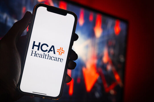 Hand Holding Phone with HCA Healthcare (HCA) Logo and Monitor in the Background Showing Red Arrows Pointing Down Suggesting Stock Decrease