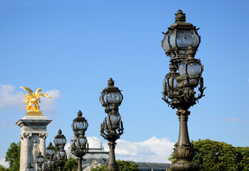 Beautiful forging lamps and golden statues of Alexander III Bridge under blue sky. Paris, France. Petit palais museum roof at background. Parisian sightseeing background.