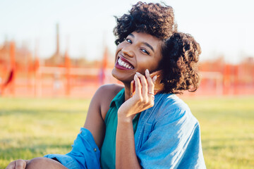 Fototapeta premium Portrait of cheerful dark skinned woman with curly hair smiling during conversation on mobile phone outdoors, attractive african american hipster girl making call sitting on green grass in park