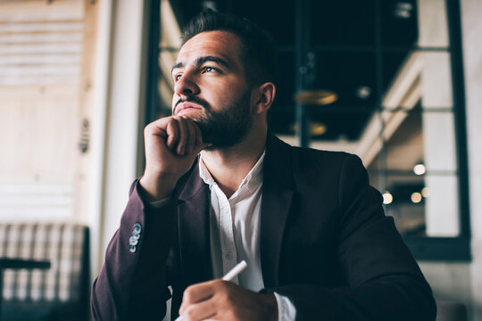 Dreamy caucasian bearded business owner looking away thinking about future working in his office, pensive formally dressed male entrepreneur pondering on successful strategy making notes in notepad