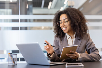 African American young woman sitting at a desk indoors, holding a notebook and a pen, talking and explaining on a video call of a laptop