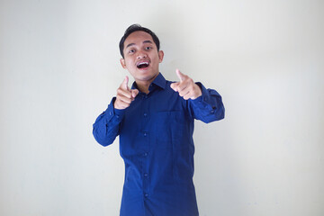 Cheerful young asian man in blue shirt pointing directly at the camera and up, expressing joy and positivity on plain background, ideal for motivational and promotional content on white background.
