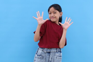 Happy asian little girl standing while showing ten fingers. Isolated on blue