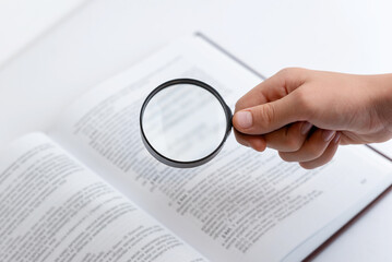 Hand holding magnifying glass searching through open book with dense text, symbolizing research,...