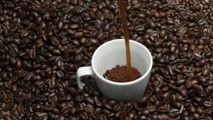 Freshly espresso was poured in white cup with pile of coffee bean. Pouring black coffee in the cup with brown background and seed scattering around on wooden table. Close up. Top view. Comestible.