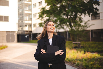 Woman entrepreneur with crossed hands hold tablet is standing on office building background. Successful smiling caucasian woman in business suit. Female worker with crossed arms look happy.