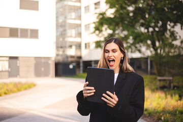 Fototapeta premium Happy delighted satisfied business woman looking tablet gesturing yes winning wow pose standing at urban city office. Overjoyed excited girl in jacket doing winner gesture, read good news.