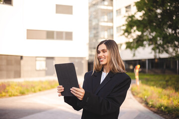 Fototapeta premium Smiling professional business woman bank manager, happy female executive or lady entrepreneur holding digital tablet pad standing near office at work on the street. Girl in suit read news, messages. 