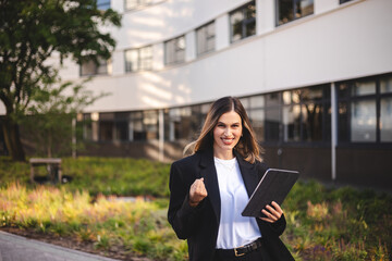 Happy delighted satisfied business woman looking tablet gesturing yes winning wow pose standing at urban city office. Overjoyed excited girl in jacket doing winner gesture, read good news.