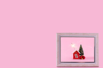 A small house with a sparkler, Christmas tree, and red ball decorations in a picture frame on a pastel pink background. Happy Christmas idea and holiday celebration concept. Copy space.