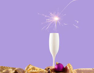 Champagne glass with a sparkler, shiny glittering decoration ball and golden metallic glowing sequins textile on pastel purple background. Glamorous holiday celebration idea. Copy space. 