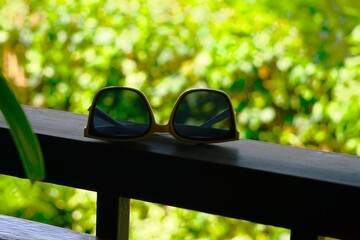 Okulary przeciwsłoneczne postawione na ogrodzeniu. Słoneczne lato i przyciemnione okulary widoczne z bliskiej odległości. Okulary przeciwsłoneczne na pierwszym planie, w tle widać zieleń. © Adam