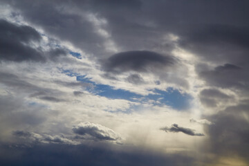 Streaks of light and blue sky break through gray clouds, variable cloudiness, overcast weather, November sky, heavenly background, celestial landscape.