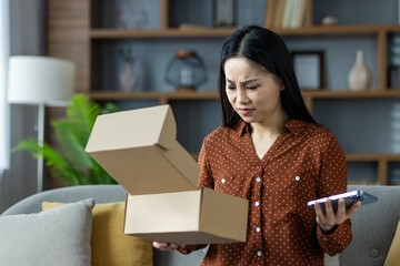Asian woman holding phone, opening box with confused expression at home. Highlights online shopping mishap or delivery issue, showcasing customer dissatisfaction and unexpected packaging.