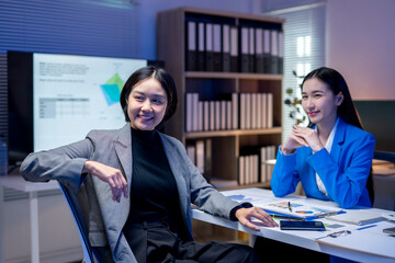 Two businesswomen smiling and discussing work in office meeting room at night