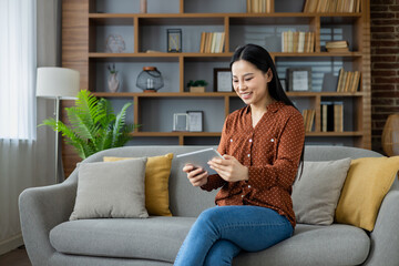 Obraz premium Asian woman plays games on tablet while sitting comfortably on sofa in cozy living room. Enjoys relaxed leisure time, digital interaction, and fun gaming experience.