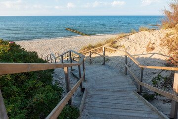 Wooden stairs descend from a sand dune to the beach. Sky, horizon and sea with waves and breakwater are visible. Background. Landscape.