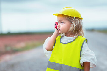 young girl wearing a yellow vest and a hard hat is talking on a cell phone