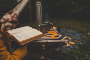 A cozy autumn moment by a campfire, featuring a person holding a warm drink in a metal mug and an...