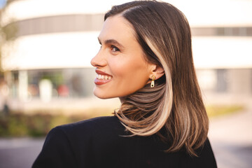 Shot of fashionable successful blonde woman in formal black jacket turning head with welcoming friendly smile, standing outdoor in city street. People, career and employment. 