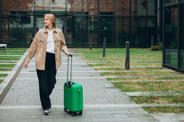 Cheerful young woman in casual outfit walking on the city park with green luggage. 30s Woman...