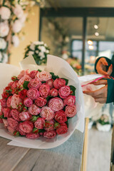 Elegant bouquet of pink roses wrapped in white paper with soft pink ribbons on a wooden table. A florist in the background holding the arrangement, highlighting the craftsmanship of flower design