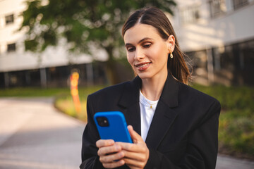 Fototapeta premium Young adult smiling professional short hair business woman wearing suit holding smartphone looking at mobile phone using apps on cellphone technology texting standing on urban city street outside.
