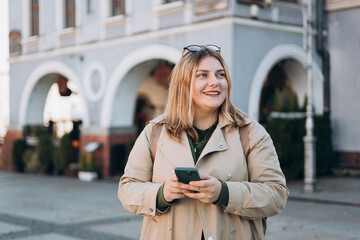 Happy cheerful young woman with backpack walking on city street checks her smartphone. Portrait of beautiful 30s girl using smartphone outdoors. Urban lifestyle concept.