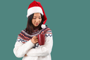 Young beautiful Asian woman in Santa hat with cup of tea on green background