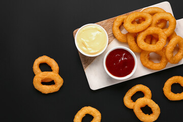 Board with fried breaded onion rings and different sauces on black background