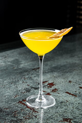 A vibrant yellow cocktail in a classic martini glass, garnished with a twist of citrus peel, set against a dark background with a textured surface. Perfect for a and elegant drink presentation.