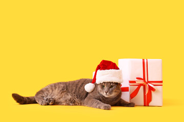 Cute cat in Santa hat with Christmas presents lying on yellow background