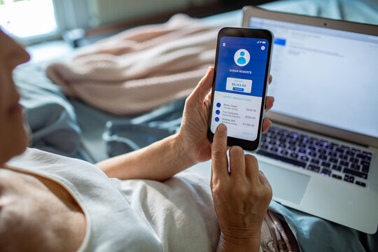 Senior woman managing finances through mobile banking app and laptop in bed