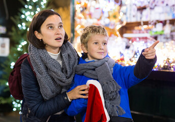 Cheerful tween boy having fun at Christmas street market with his mother, looking in amazement at...