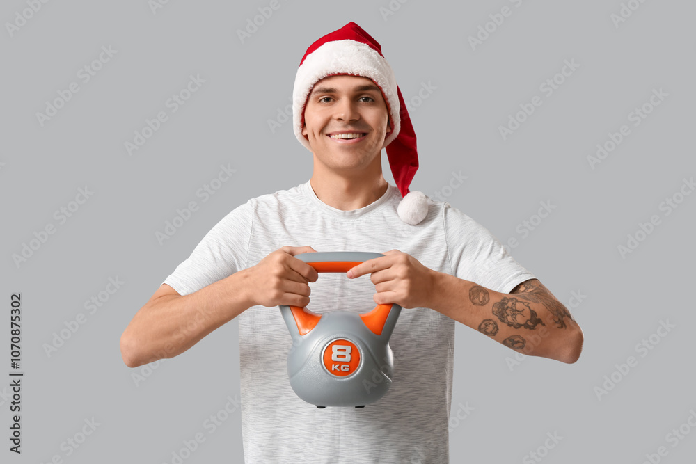 Poster Young man in Santa hat training with kettlebell on grey background