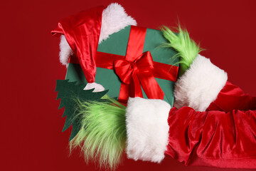 Green hairy hands of creature in costume with Christmas gift box and Santa hat on red background