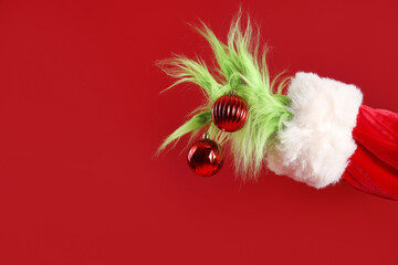 Obraz premium Green hairy hand of creature in Santa costume with Christmas balls on red background