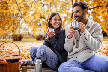Young happy couple having picnic in a grass in a colorful autumn park. They drink coffee from thermos.