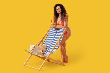 Beautiful young African-American woman with glass of cocktail, wicker bag and deck chair on yellow...