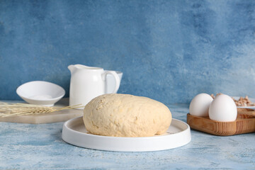 Plate with fresh raw dough, wheat and wooden holder of eggs on blue background