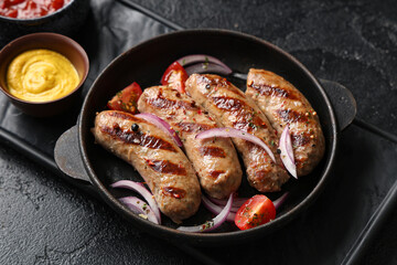 Frying pan with tasty grilled sausages, tomatoes, onion and bowl of mustard on black background