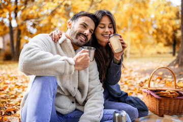 Young happy couple having picnic in a grass in a colorful autumn park. They drink coffee from thermos.