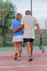 vertical senior tennis couple, seen from behind, walking off court together after a loss