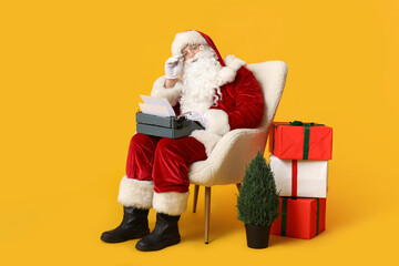 Santa Claus with vintage typewriter sitting in armchair and gift boxes on yellow background
