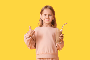 Cute little girl with glass of sweet chocolate milk showing thumb-up gesture on yellow background